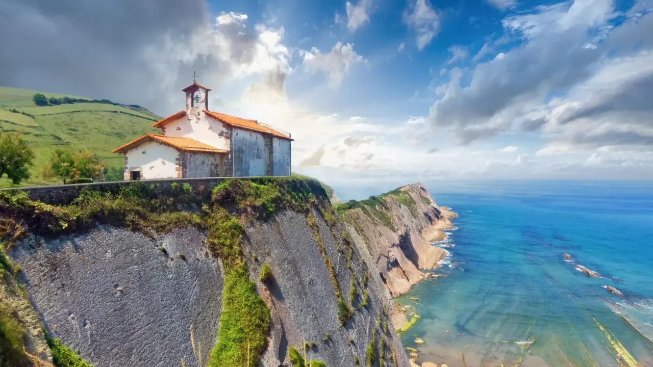 Ermita sobre impresionantes acantilados en España, con el mar azul y un cielo nublado.
