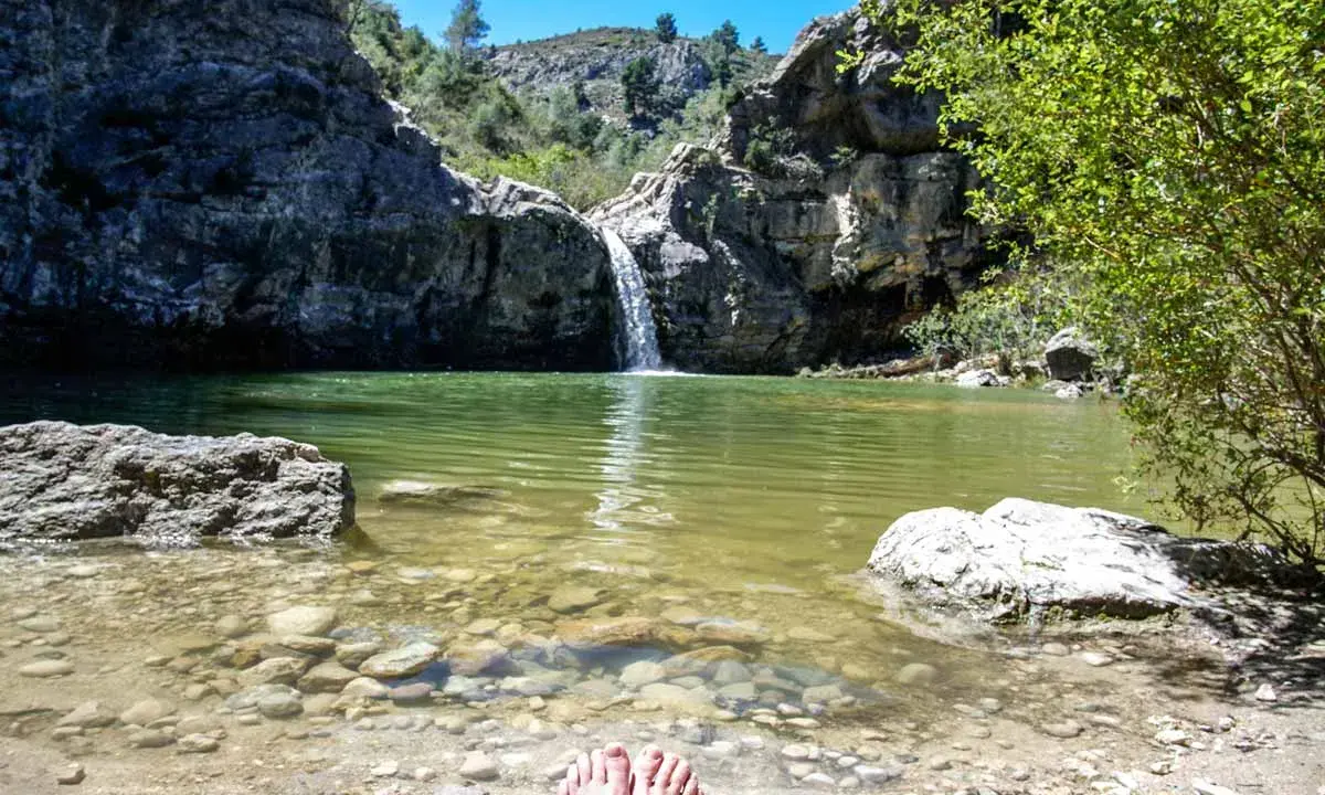 Pies descalzos en la orilla del **barranco de la Encantada**, con una cascada cayendo en un estanque de aguas verdes rodeado de rocas y vegetación.