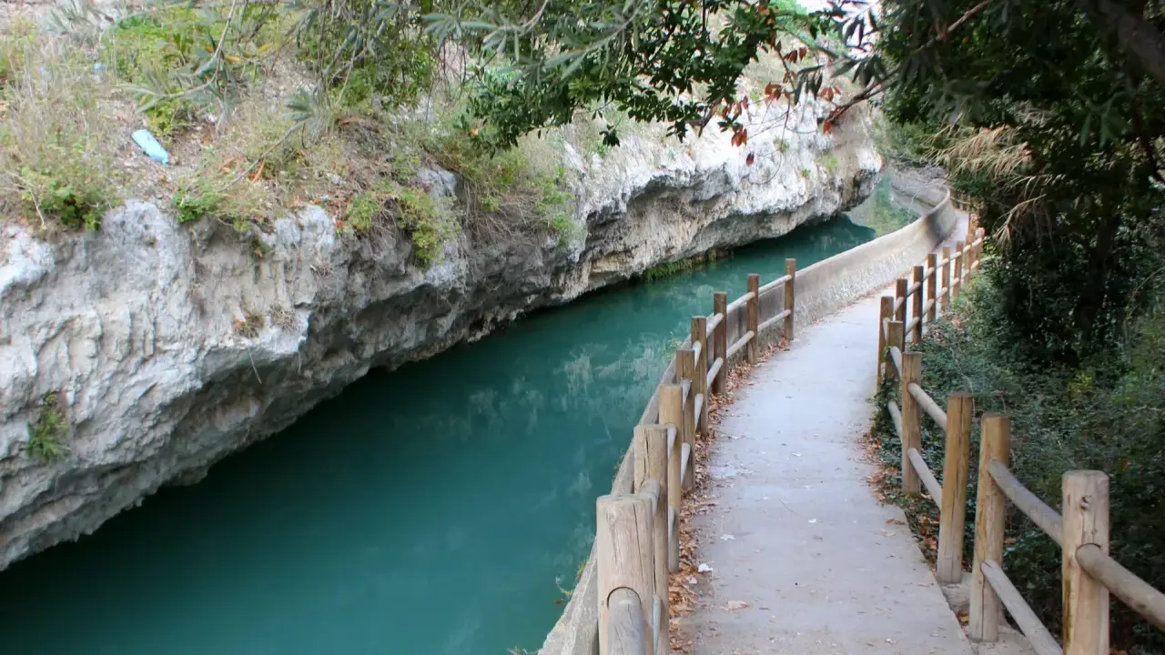 Sendero junto al río de aguas turquesas en Alcalá del Júcar, con rocas escarpadas y vegetación frondosa.