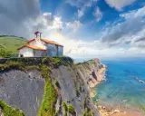 Ermita sobre impresionantes acantilados en España, con el mar azul y un cielo nublado.