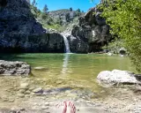 Pies descalzos en la orilla del **barranco de la Encantada**, con una cascada cayendo en un estanque de aguas verdes rodeado de rocas y vegetación.