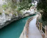 Sendero junto al río de aguas turquesas en Alcalá del Júcar, con rocas escarpadas y vegetación frondosa.