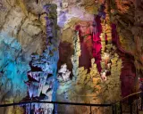 Interior de las **Canelobre caves**, con estalactitas y estalagmitas iluminadas con luces de colores azul, rosa y amarillo. Un visitante observa la maravilla geológica.
