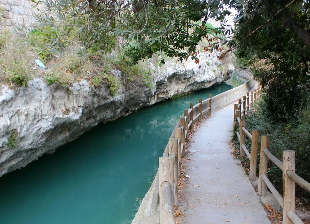 Sendero junto al río de aguas turquesas en Alcalá del Júcar, con rocas escarpadas y vegetación frondosa.