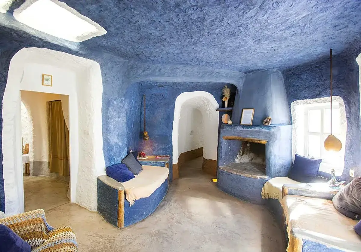 Interior de una acogedora casa cueva con paredes y techo azul texturizado, chimenea, sofás curvos y una claraboya.