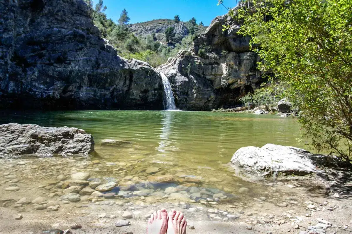 Pies descalzos en la orilla del barranco de la Encantada, con una cascada cayendo en un estanque de aguas verdes rodeado de rocas y vegetación.