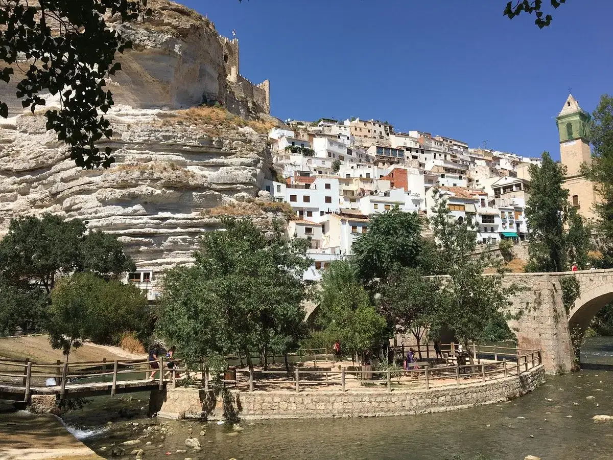 Casas blancas se aferran a un acantilado junto al río Alcalá del Júcar. Un puente de piedra cruza el agua.