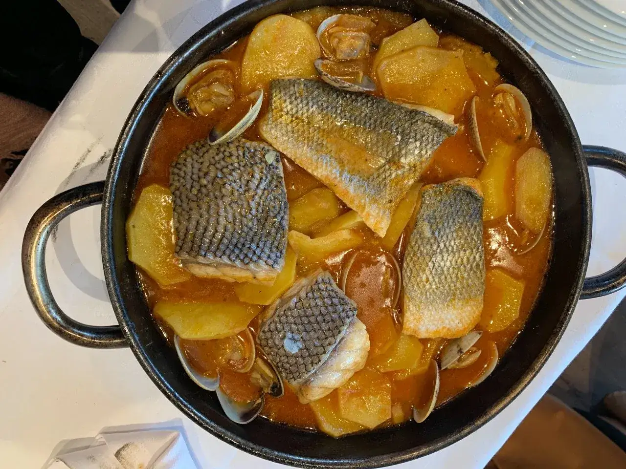 Delicioso plato de comida gallega: trozos de pescado con piel, patatas y almejas en una salsa rojiza, servido en una cazuela negra.