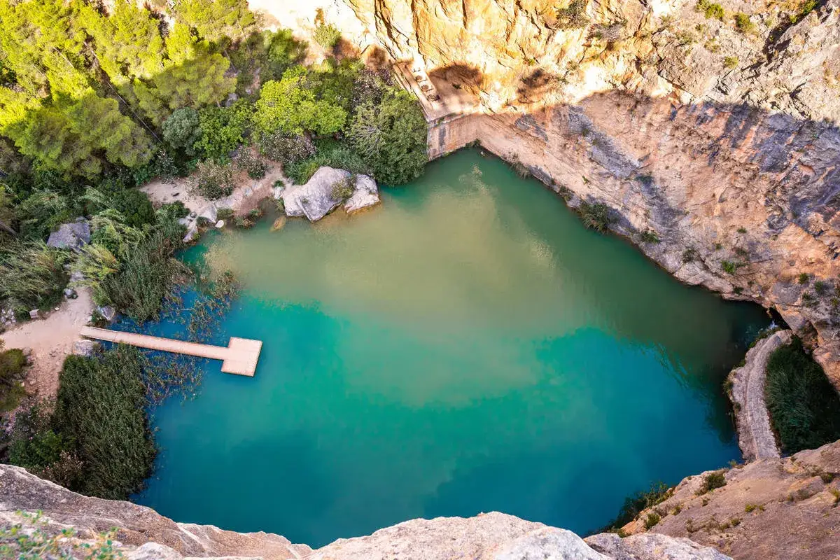 Un charco azul en Chiva, rodeado de rocas y vegetaci&oacute;n. Un muelle de madera se adentra en sus aguas cristalinas.