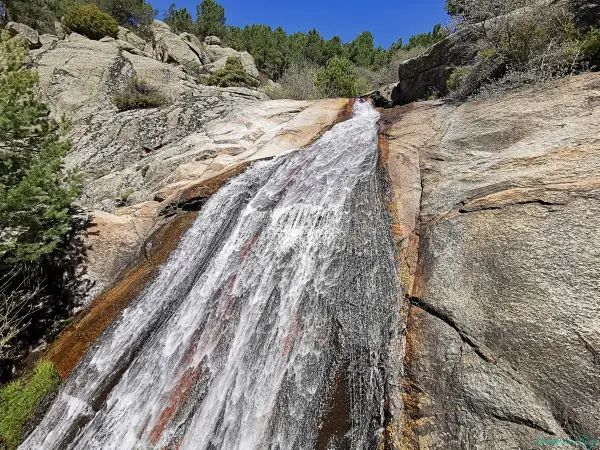 El agua desciende por la roca en la cascada del Hornillo, rodeada de vegetación y rocas graníticas bajo un cielo azul.