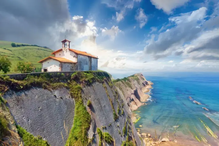 Ermita sobre impresionantes acantilados en España, con el mar azul y un cielo nublado.