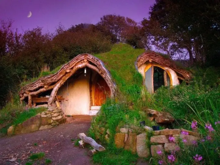 Encantadora casa cueva con techos de madera y césped, integrada en una colina bajo un cielo púrpura con luna creciente.