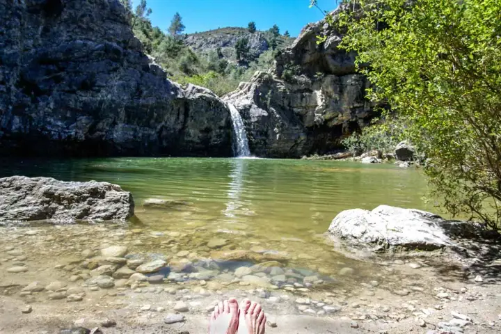 Pies descalzos en la orilla del **barranco de la Encantada**, con una cascada cayendo en un estanque de aguas verdes rodeado de rocas y vegetación.