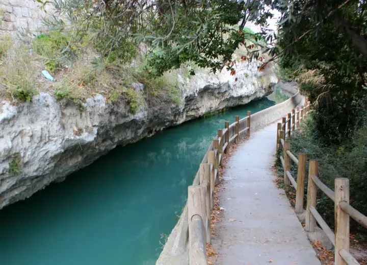 Sendero junto al río de aguas turquesas en Alcalá del Júcar, con rocas escarpadas y vegetación frondosa.