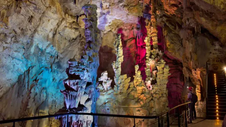 Interior de las **Canelobre caves**, con estalactitas y estalagmitas iluminadas con luces de colores azul, rosa y amarillo. Un visitante observa la maravilla geológica.